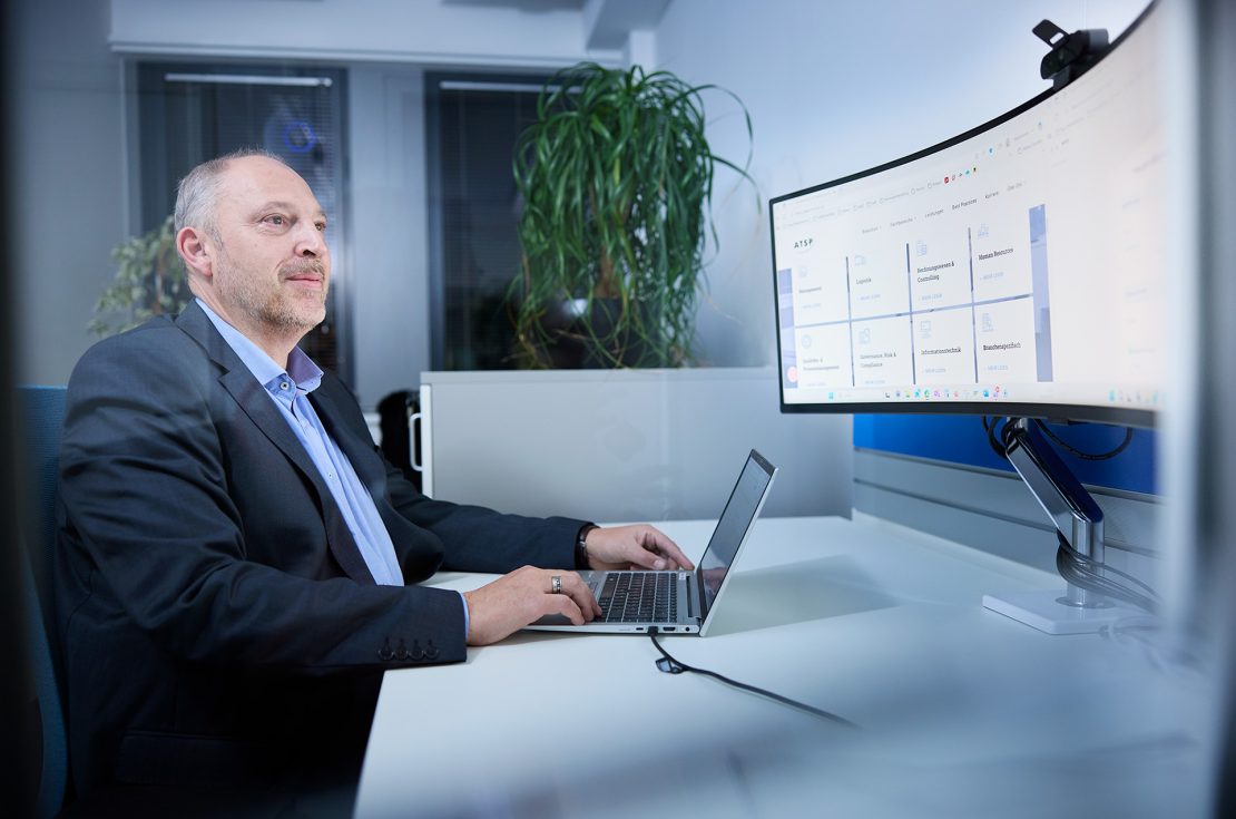 Mann im Anzug sitzt am Schreibtisch mit Laptop und Monitor